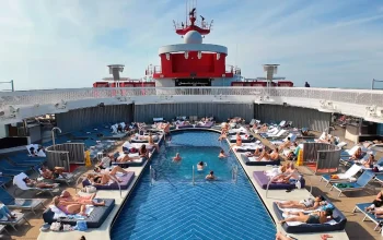 A lively view of the Virgin Voyages cruise ship Scarlet Lady’s pool deck, featuring people playing in the water, dancing, and socializing under sunny skies, with loungers and the ship’s modern design in the background.