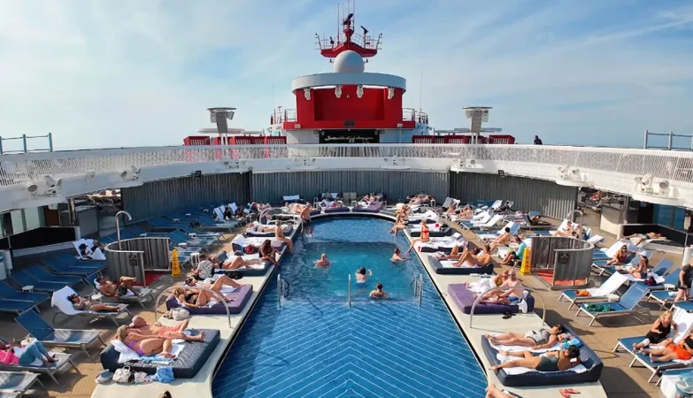 A lively view of the Virgin Voyages cruise ship Scarlet Lady’s pool deck, featuring people playing in the water, dancing, and socializing under sunny skies, with loungers and the ship’s modern design in the background.